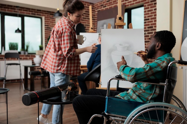 Leraar legt illustratieproces uit aan verlamde student met een handicap die werkt aan een schilderproject tijdens artistieke les in creativiteitsstudio. Divers team dat vaas schetst met grafisch potlood