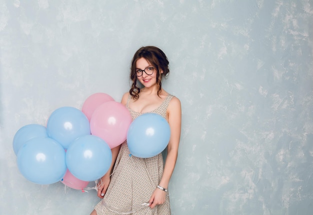 Leuk donkerbruin meisje dat zich in een studio bevindt, wijd glimlacht en blauwe en roze ballonnen vasthoudt. Ze draagt een licht gestippelde jurk en een zwarte bril, en heeft gevlochten haar.