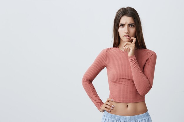 Leuk knap jong Kaukasisch meisje met lang bruin haar in de modieuze hand van de uitrustingsholding dichtbij mond met onzekere en ongerust gemaakte uitdrukking terwijl het luisteren van examen resulteert in universiteit.