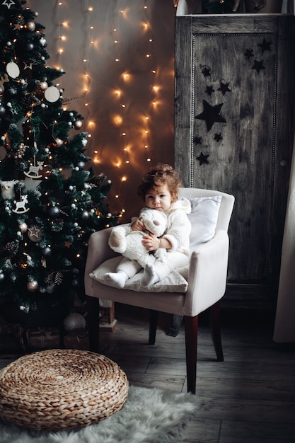 Leuk krullend kind met een pluche beer zittend in een fauteuil bij een prachtig versierde kerstboom