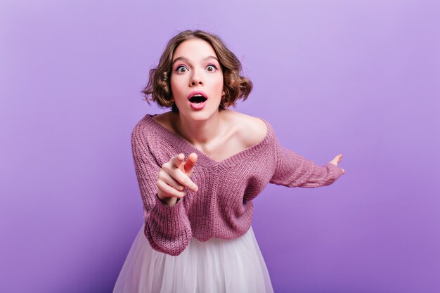 Leuk meisje dat met roze make-up verbazing en wijzende vinger uitdrukt. Binnenfoto van gekrulde geschokte vrouw die op purpere muur wordt geïsoleerd.