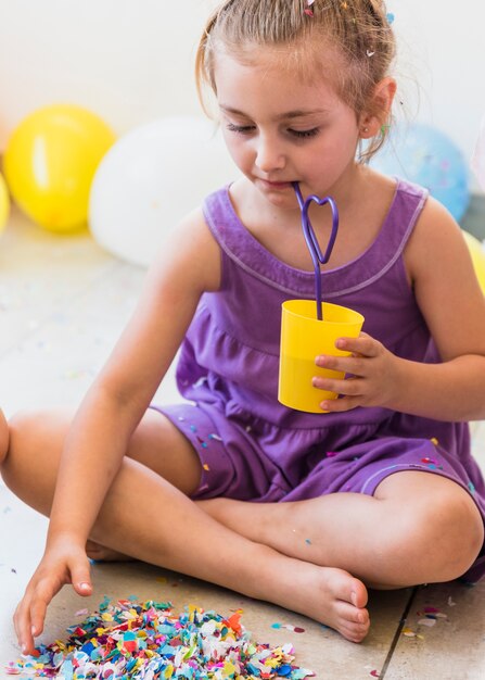 Leuk meisje die sap met stro drinken die confettien op vloer verzamelen