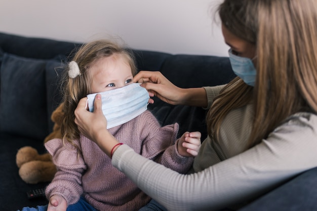 Leuk meisje en moeder die gezichtsmasker dragen, thuis zittend op bed, die droevige peuterdochter troosten