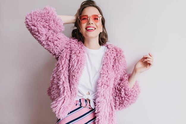 Leuk meisje in roze zonnebril poseren met vrolijke glimlach op witte muur. Indoor portret van kortharige vrouw in bontjas spelen met haar haar.