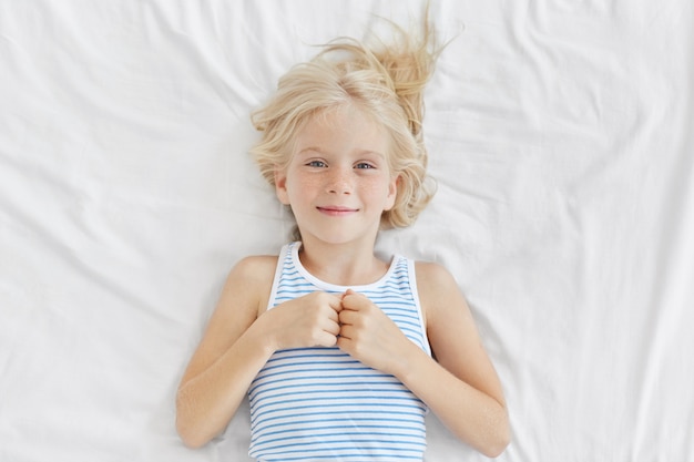 Leuk meisje met blond haar, blauwe charmante ogen en sproetengezicht, zeeman T-shirt dragend, liggend op witte beddekking, blije uitdrukking na prettige dromen 's nachts. Kinderen, ontspanning