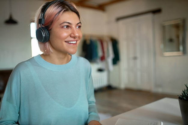 Leuk meisje met neusring en roze haren zit aan bureau in draadloze hoofdtelefoons, met stemles met behulp van webcam videochat, online leren, opgewonden vrolijke blik hebben. mensen en technologie