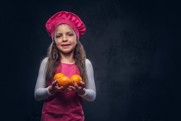 Leuk mooi schoolmeisje gekleed in een roze koksuniform houdt sinaasappels vast in een studio. Geïsoleerd op een donkere gestructureerde achtergrond.