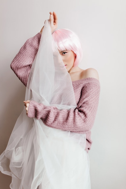 Leuk prachtig meisje met roze haar verbergend gezicht achter witte doek. glamoureus vrouwelijk model in glanzende periwig speels poseren op lichte muur.