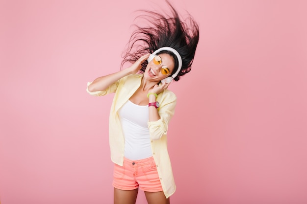 Leuk spaans meisje met gebruinde huid trendy armband en oranje bril luisteren muziek en dansen. indoor foto van aantrekkelijke latijnse dame in geel katoenen jasje met plezier.