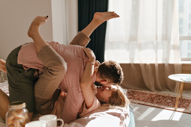 Leuk stel in pastelkleurige kleren ligt op bed en is in een goed humeur aan het dollen in lichte appartementen.