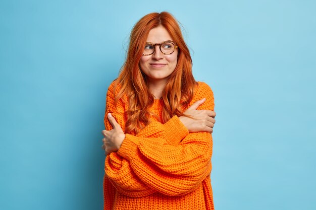 Leuk uitziende roodharige vrouw omhelst zichzelf graag dat haar nieuwe gebreide oranje trui er aan de rechterkant tevreden uitziet.