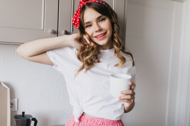 Leuk wit meisje in casual t-shirt staan in de keuken met een kopje thee. Binnen schot van ontspannen vrouwelijk model koffie drinken in de ochtend.