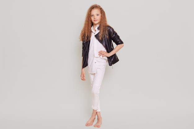 Leuk zelfverzekerd schoolmeisje die witte blouse, broek en leerjasje dragen die zich met hand op heup bevinden, die voorzijde bekijken