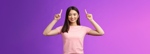 Leuke assertieve donkerharige Aziatische vrouw draagt een roze t-shirt en steekt de handen omhoog met promo wijzende vingers