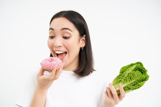 Leuke aziatische vrouw slaat dieet over en neemt hap van heerlijke geglazuurde donut die over witte achtergrond staat
