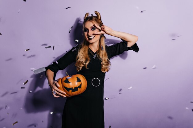 Leuke blanke vrouw in zwarte jurk poseren na halloween-maskerade. Binnenfoto van glimlachend vrolijk meisje met pompoen.