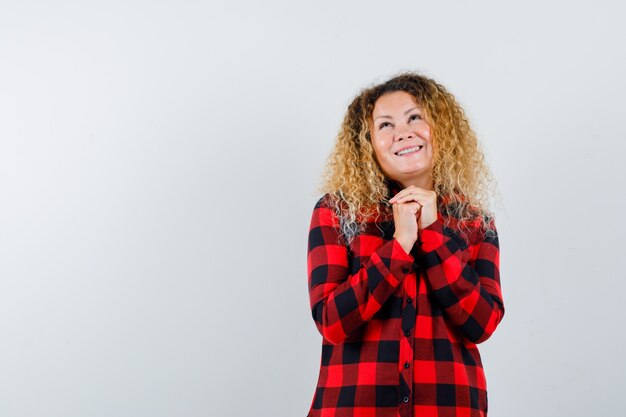 Leuke blonde vrouw die haar handen vasthoudt in een smekend gebaar in een geruit overhemd en er hoopvol uitziet. vooraanzicht.