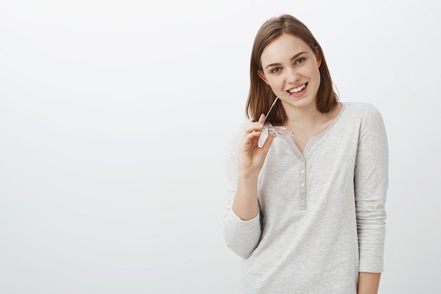 Leuke collega hebben en oog op charmante kerel flirten tijdens pauze bijten brillen brillen in de hand houden glimlachend teder poseren tegen grijze muur in casual witte blouse