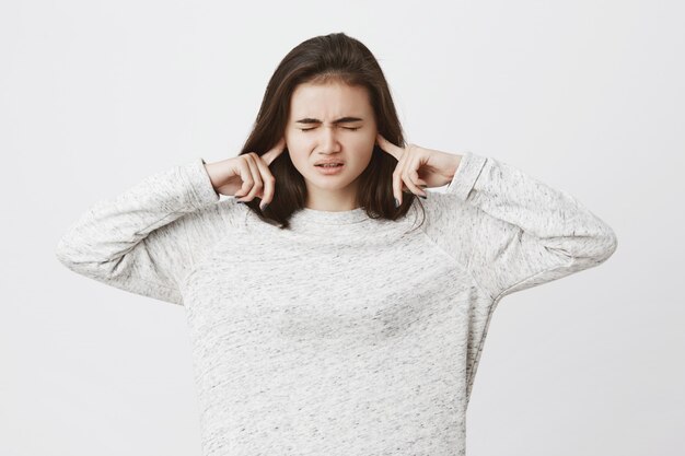 leuke donkerbruine vrouw, die spanning en druk uitdrukt, vingers in oren houdt om iets niet te horen en haar ogen sluit