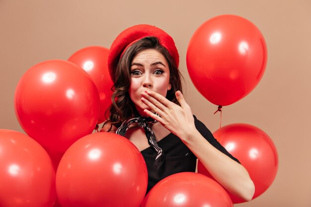 Leuke donkerharige dame kijkt gelukkig naar de camera en blaast kus op beige achtergrond met rode ballonnen.