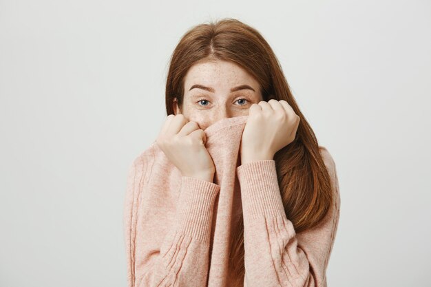 Leuke dwaze roodharige vrouw verbergt gezicht achter trui kraag en ziet er verlegen uit