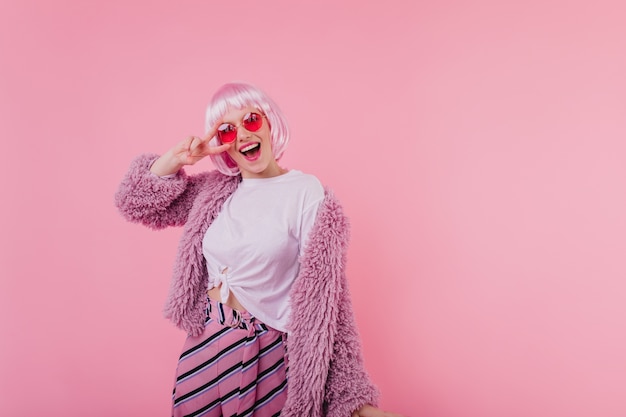 Leuke emotionele dame in roze korte periwig die voor de gek houdt. Foto van gelukkig Europees vrouwelijk model in zonnebril en pluizig jasje dat op heldere muur wordt geïsoleerd