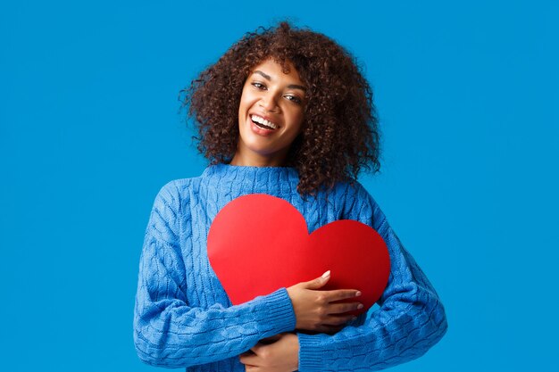 Leuke en tedere grappige, glimlachende Afro-Amerikaanse vrouw met afro-kapsel, druk het grote rode hartteken tegen de borst en omhels het met een opgetogen charmante grijns, met liefde en genegenheid, blauwe muur.