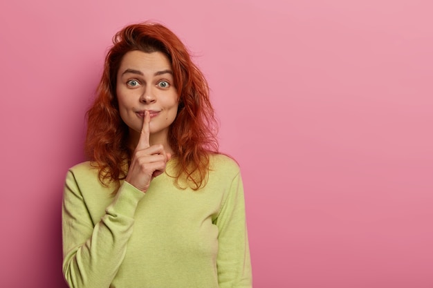 Gratis foto leuke enthousiaste vrouw met rood golvend haar, roddelen en vragen geen geruchten verspreiden, houdt de vinger op de lippen gedrukt