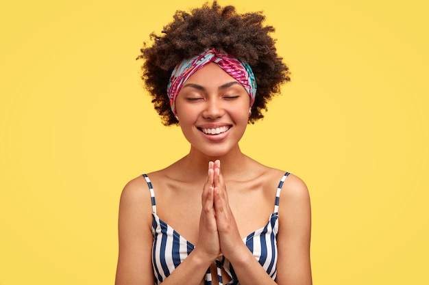 Gratis foto leuke gelukkige afro-amerikaanse vrouw houdt de handen in het bidden gebaar, heeft een brede glimlach, bidt voor een belangrijke gebeurtenis, gekleed in een gestreept t-shirt, vormt tegen gele muur. geloof in beter