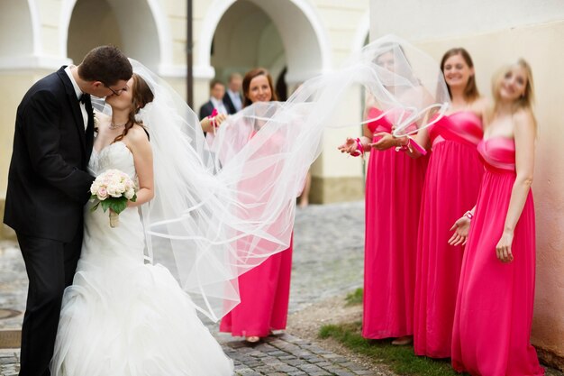 Leuke gelukkige jonge bruidegom en bruid kussen op de achtergrond getuigen in de roze jurken