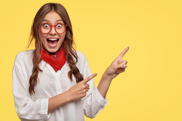 Leuke gelukkige vrouw toont de weg, geeft richting, wijst met beide handen naar rechts, heeft dolgelukkig gelaatsuitdrukking, draagt stijlvolle kleding, modellen over gele muur. Advertentie concept