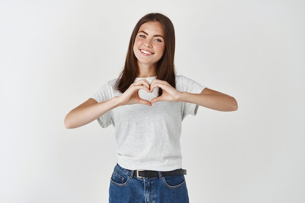 Leuke jonge vrouw die een hartteken toont, ik hou van je gebaar, ik wens een gelukkige Valentijnsdag, staande over een witte muur.