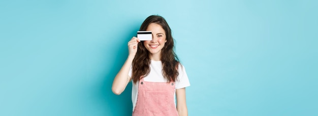 Leuke jonge vrouw die een plastic creditcard op het oog laat zien en glimlachend winkelend terwijl het over blauw staat