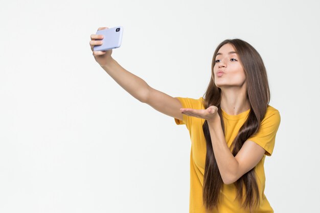 Leuke jonge vrouw die een selfie nemen en lucht-kus geven die op witte muur wordt geïsoleerd