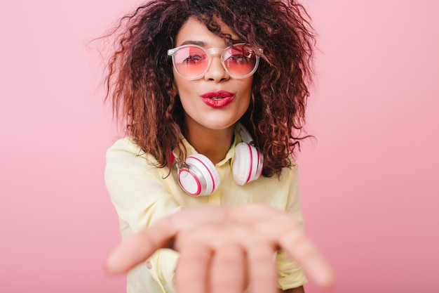 Leuke jonge vrouw met Afrikaans kapsel die luchtkus verzendt en met mooie gezichtsuitdrukking poseert. Charmante mulat dame in elegant geel shirt en witte koptelefoon met plezier op roze achtergrond.