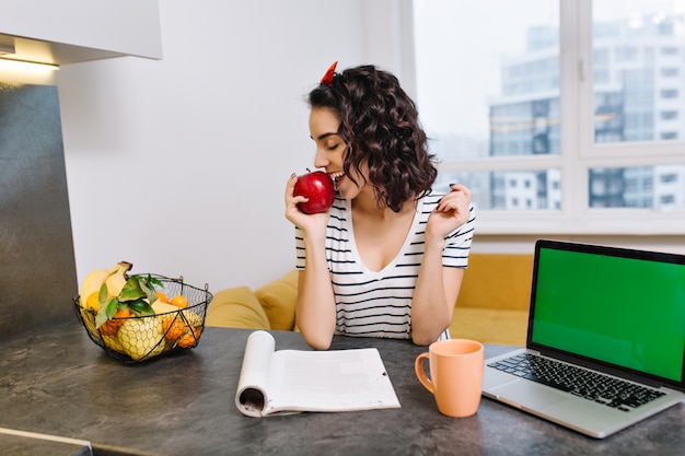 Leuke jonge vrouw met krullend geknipt haar op tafel in modern appartement. Rode appel eten, lachend met gesloten ogen, werkplek thuis, laptop met groen scherm, rust