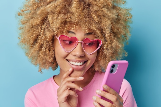 Leuke jonge vrouw met krullend haar draagt stijlvolle roze hartvormige zonnebril en t-shirt houdt mobiele telefoon chats met vriendje horloges video geïsoleerd op blauwe achtergrond Moderne technologieën