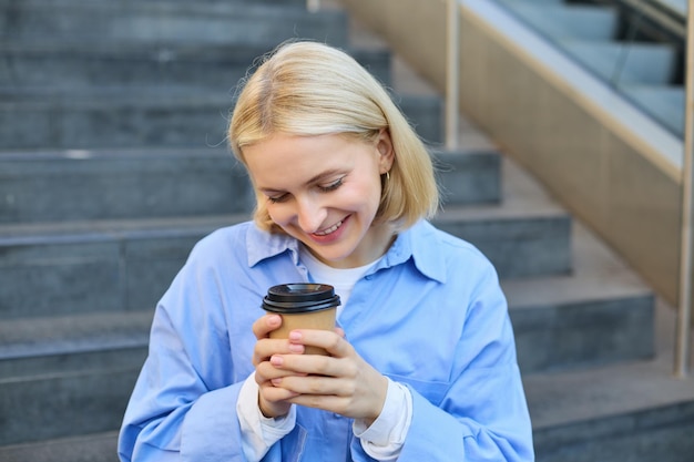 Leuke jonge vrouwelijke student vrouw met een kop koffie in de handen rustend op de trap in het centrum van de stad zit