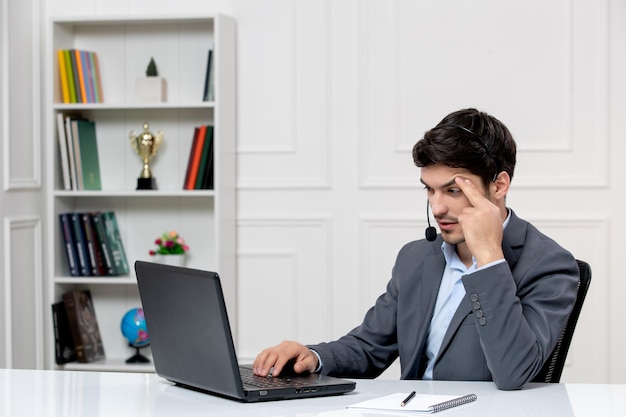 Leuke kerel van de klantenservice in grijs pak met computer en headset die voorhoofd aanraakt en moe?