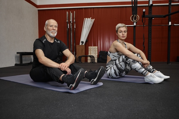 Leuke sportieve jonge vrouw met kort kapsel zittend op de vloer met aantrekkelijke bebaarde man gepensioneerde m / v in sportkleding met vermoeide gezichtsuitdrukkingen, ontspannen tussen sets van crossfit oefeningen