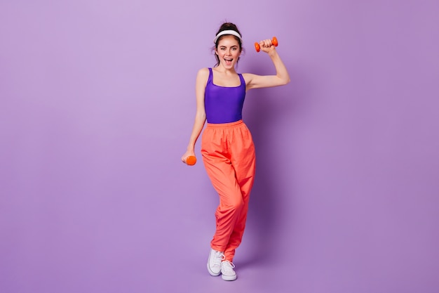 Leuke sportieve vrouw in lichte fitness outfit in stijl van de jaren 80 met glimlach toont oefeningen met halters