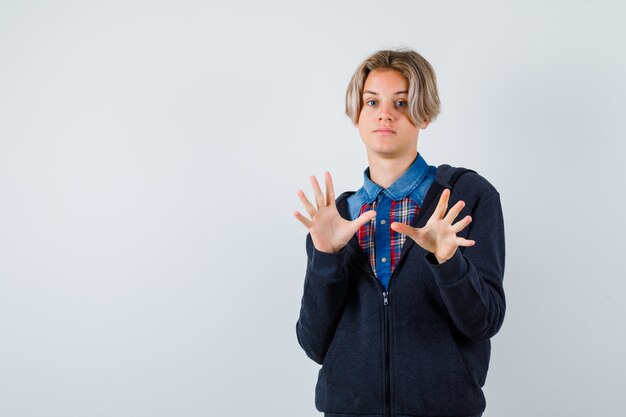 Leuke tienerjongen die overgavegebaar in overhemd, hoodie toont en terughoudend kijkt, vooraanzicht.