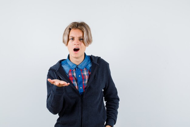 Leuke tienerjongen die palm aan de voorkant in shirt, hoodie spreidt en verbijsterd kijkt. vooraanzicht.