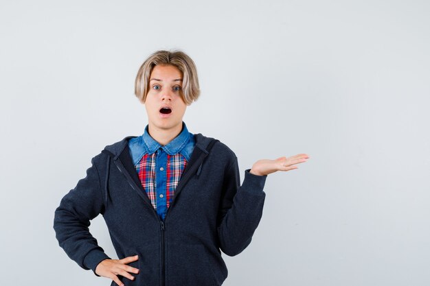 Leuke tienerjongen die palm opzij spreidt in shirt, hoodie en verrast kijkt. vooraanzicht.