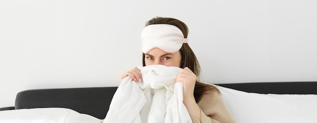Gratis foto leuke vriendin met slaapmasker op voorhoofd bedekt haar gezicht achter dekbed en giechelt