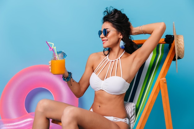 Leuke vrouw cocktail drinken en spelen met haar studio shot van vrolijk gebruind meisje in bikini zittend op chaise longue