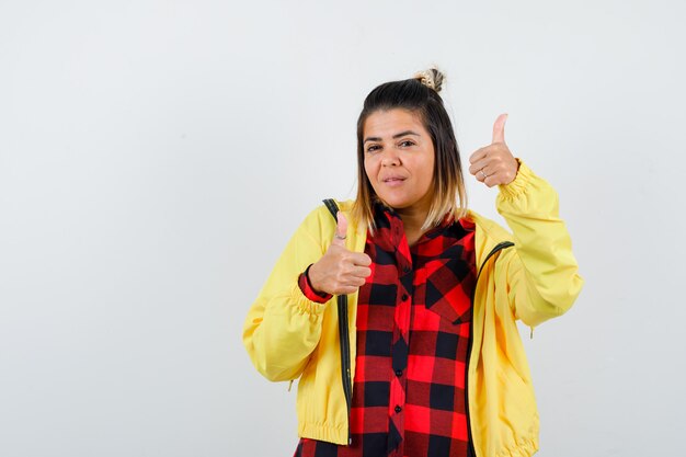 Leuke vrouw die dubbele duimen in shirt, jas toont en er zelfverzekerd uitziet, vooraanzicht.