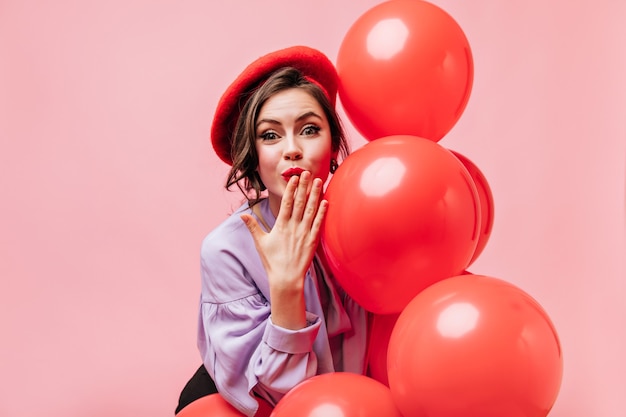 Leuke vrouw in lila blouse en rode baret blaast kus en houdt ballonnen op roze achtergrond.