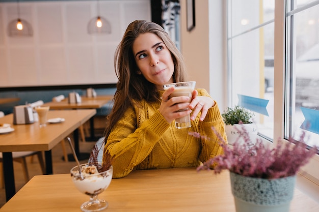Leuke vrouw in trendy gele trui iets na te denken tijdens het rusten in café met glas cappuccino. Indoor portret van prachtige dame wachten op vriend en genieten van ijs.