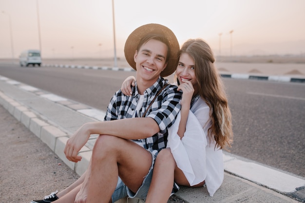 Gratis foto leuke vrouw lachen in witte blouse met plezier met vriendje in stijlvolle denim shorts, rust na een wandeling langs de weg. glimlachend mooi paar tijd samen doorbrengen met geweldige hemel